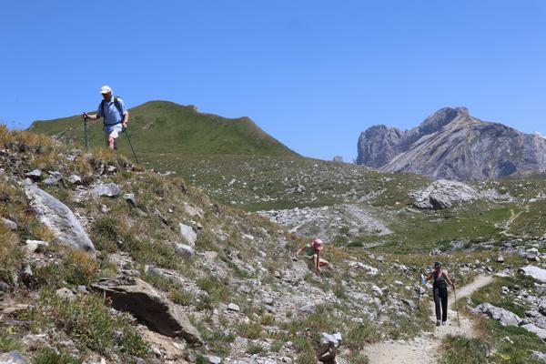 Three people, including a child, are seen hiking on a rocky mountain trail under a clear blue sky